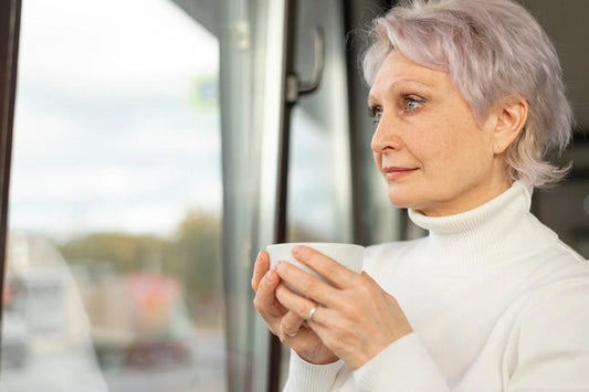Older person with gray hair and visible neck wrinkles stands near a window wearing a white shirt.